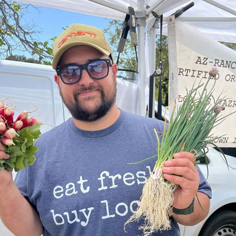 A male farmer holds spring onions and radishes. His shirt says, 